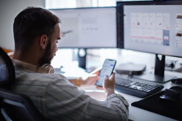 a man slouching in an office chair in front of a computer mo<em></em>nitor looks at his phone